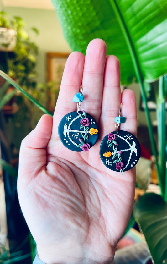 Dynamic polymer clay Sagittarius earrings featuring a bold design of a bow and arrow surrounded by hand sculpted roses. Handcrafted with precision, these earrings capture the adventurous and passionate spirit of Sagittarius. Embrace your fiery nature with this symbolic accessory. Shop now for a unique expression of Sagittarian energy!